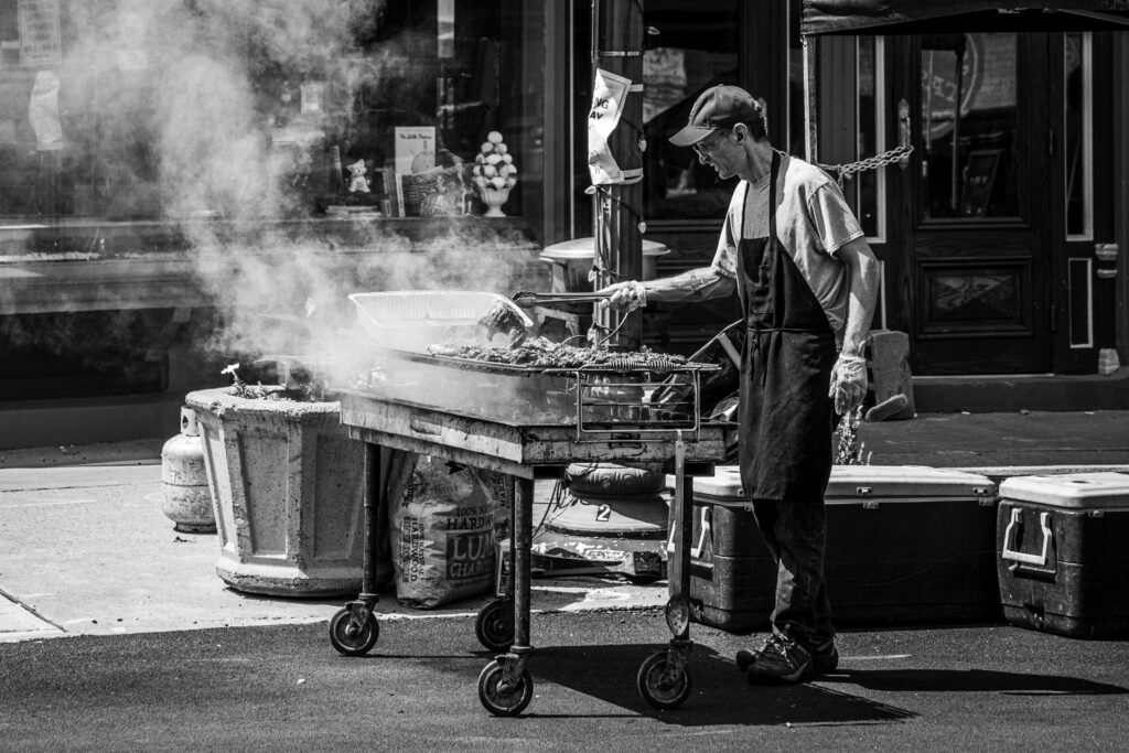 A grill master tends to a smoky barbecue on a city street, creating a vibrant urban scene.